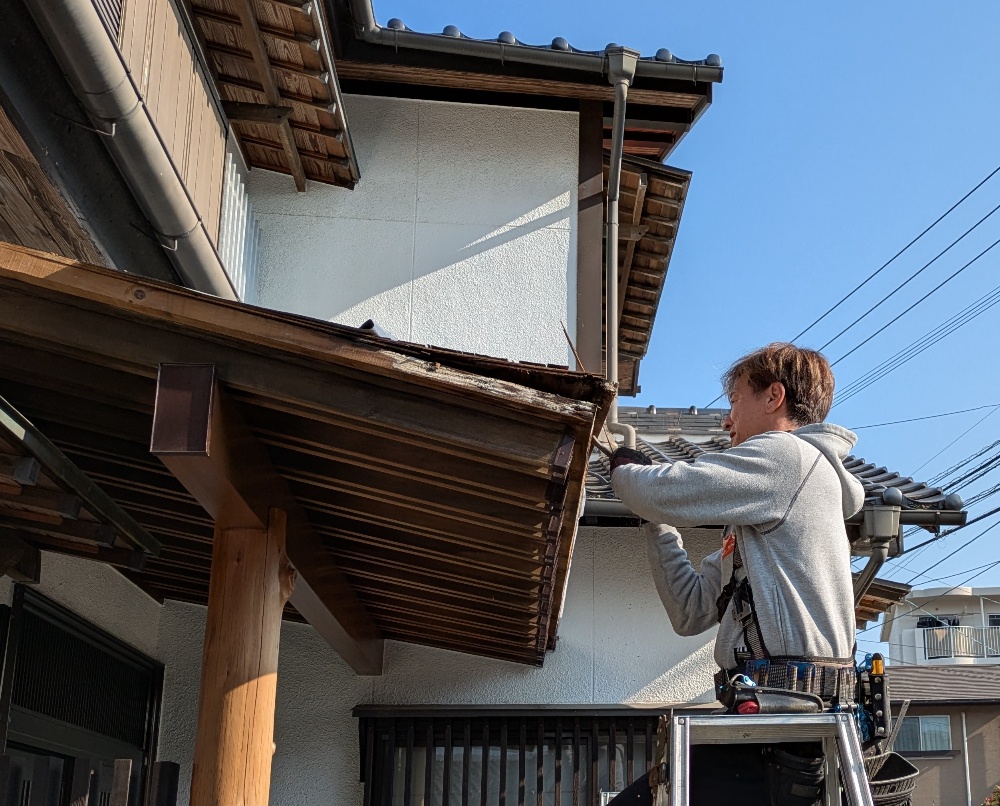 快晴の下、脚立に乗って軒樋・軒天を修繕するつながり工房のスタッフ